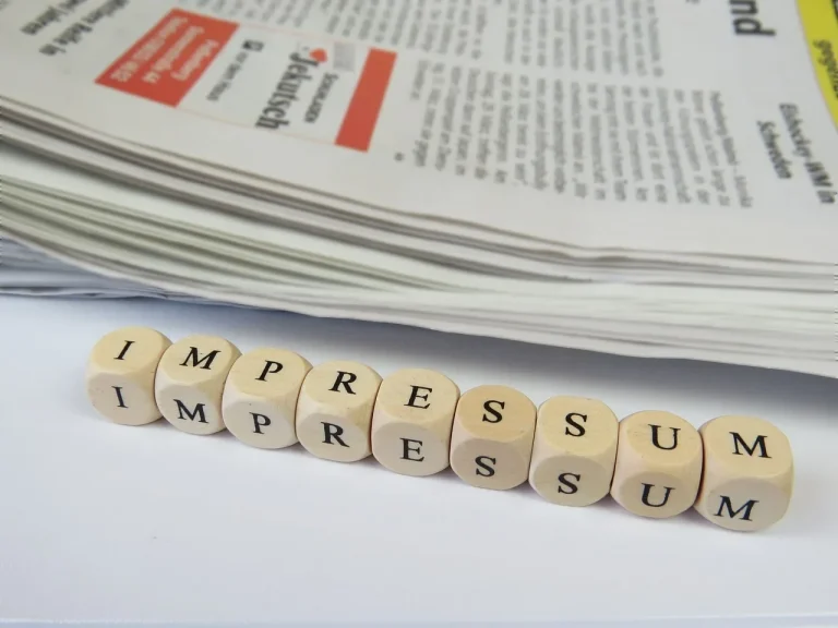 Rechtliche Transparenz: Impressum im Detail Würfel mit schwarzen Buchstaben bilden das Wort 'IMPRESSUM' in einer Reihe vor einem Stapel weißer Dokumente mit roter Markierung .